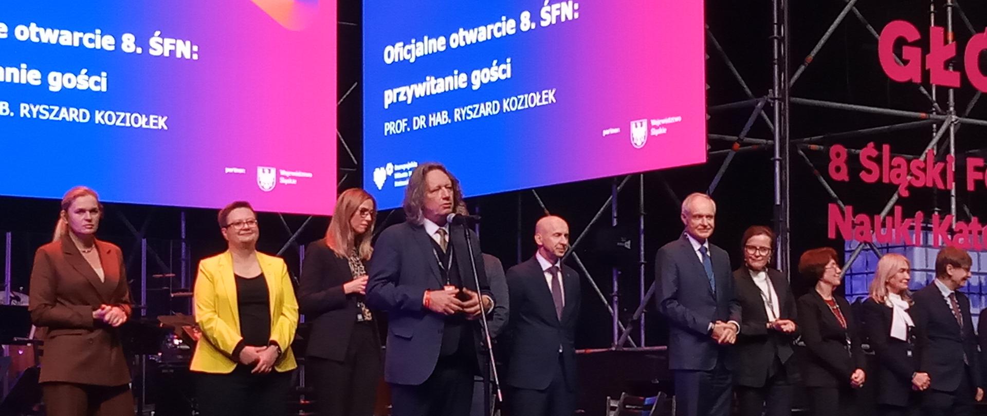 Grupa osób na scenie w światłach reflektorów, profesor Koziołek stoi przed grupą i przemawia, wita gości.