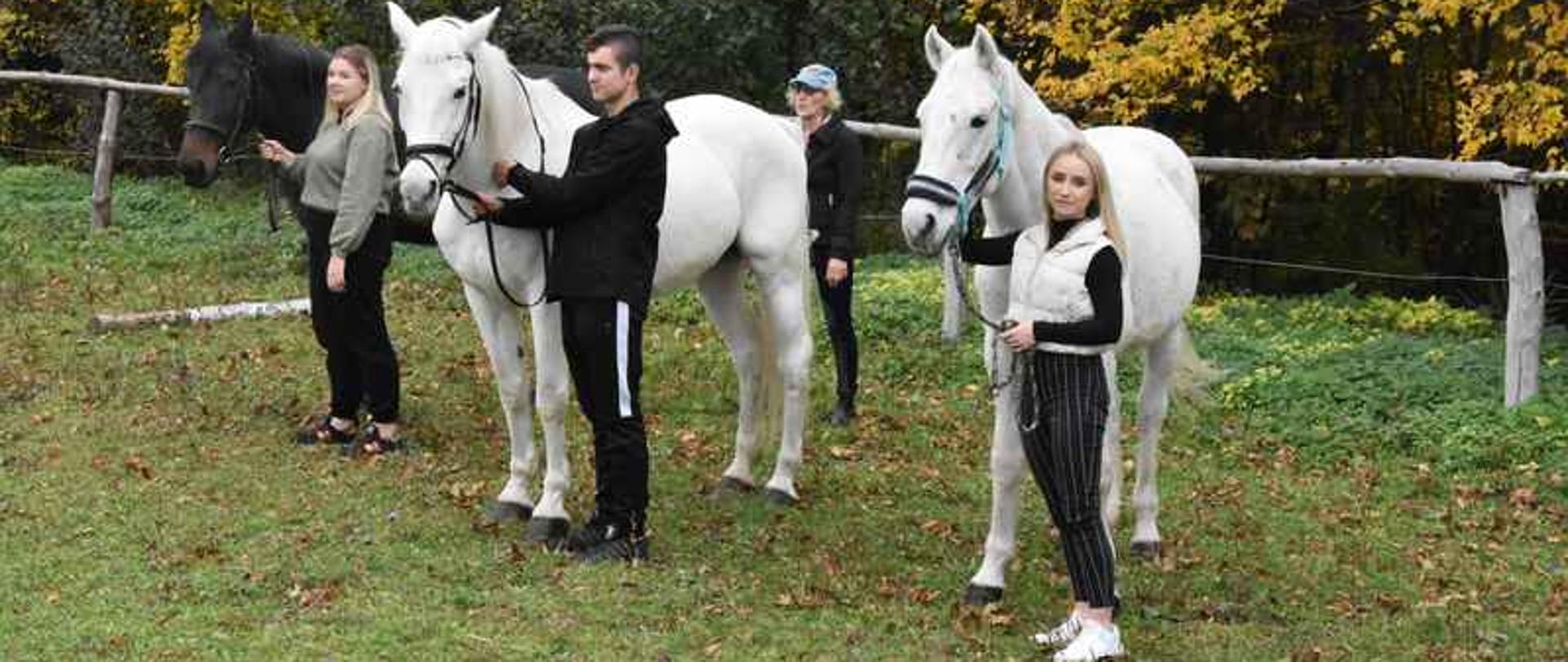 Na tle lasu stoją trzy młode osoby. Każda z osób trzyma konia.