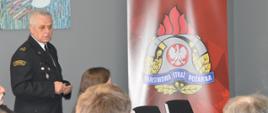 Dzień Bezpieczeństwa i Ochrony Zdrowia w Pracy. Sala konferencyjna Ferm Drobiu Woźniak w Żylicach. W niej uczestnicy konferencji - kierownicy poszczególnych działów firmy, a także przedstawiciele Komendy Powiatowej PSP w Rawiczu, Powiatowej Stacji Sanitarno-Epidemiologicznej w Rawiczu i Państwowej Inspekcji Pracy. Wykład prowadzi zastępca komendanta powiatowego PSP w Rawiczu. W lewym narożniku pomieszczenia stoi
roll-up promujący Państwową Straż Pożarną.