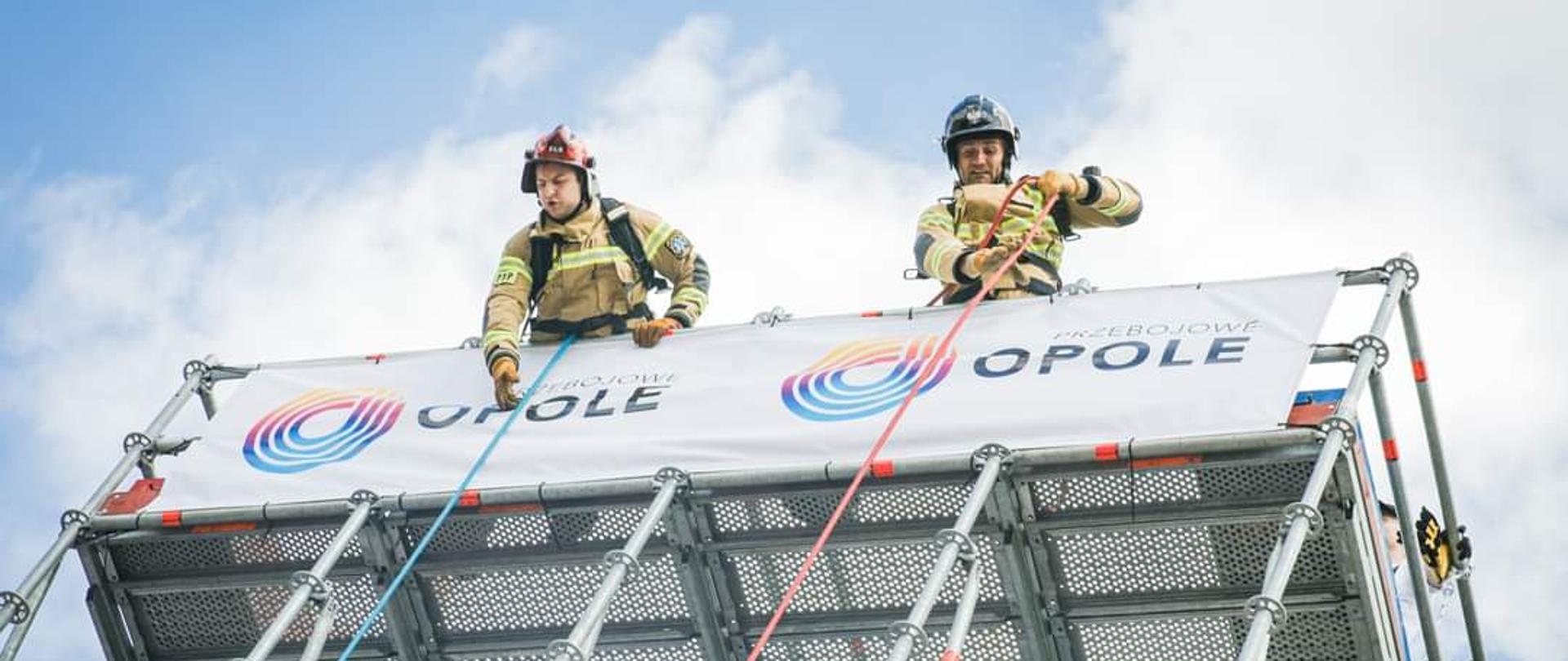 Na zdjęciu widać dwójkę zawodników na wysokości, przechylonych przez barierkę, którzy z dużym wysiłkiem wciągają do góry coś na linach. 