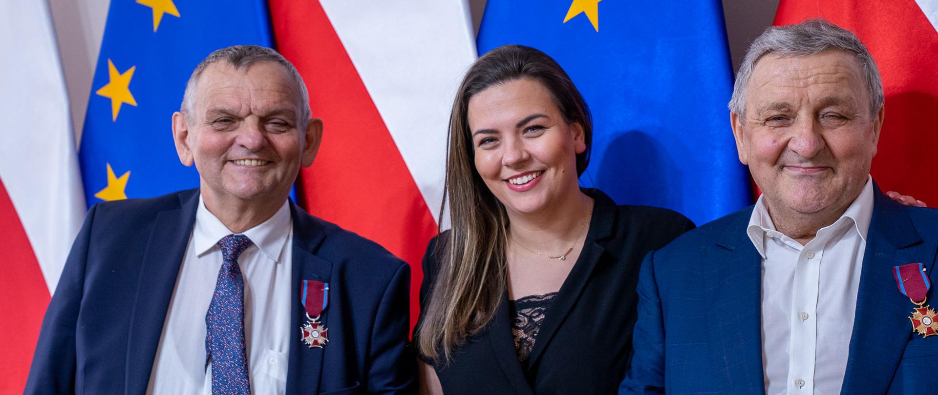 Na zdjęciu znajdują się trzy osoby, które brały udział w uroczystości. Jedna kobieta i dwóch mężczyzn, którzy stoją na tle flagi Unii i Polski 