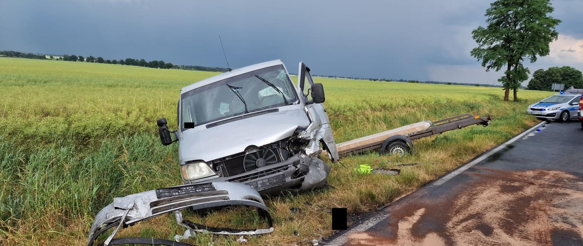 Widać srebrny samochód z przyczepa w rowie, przód pojazdu uszkodzony, za nim jest pole i granatowe niebo