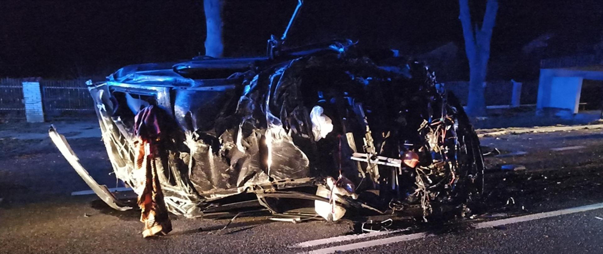 Zdjęcie przedstawia doszczętnie rozbity samochód osoby leżący na boku na pasie drogi.