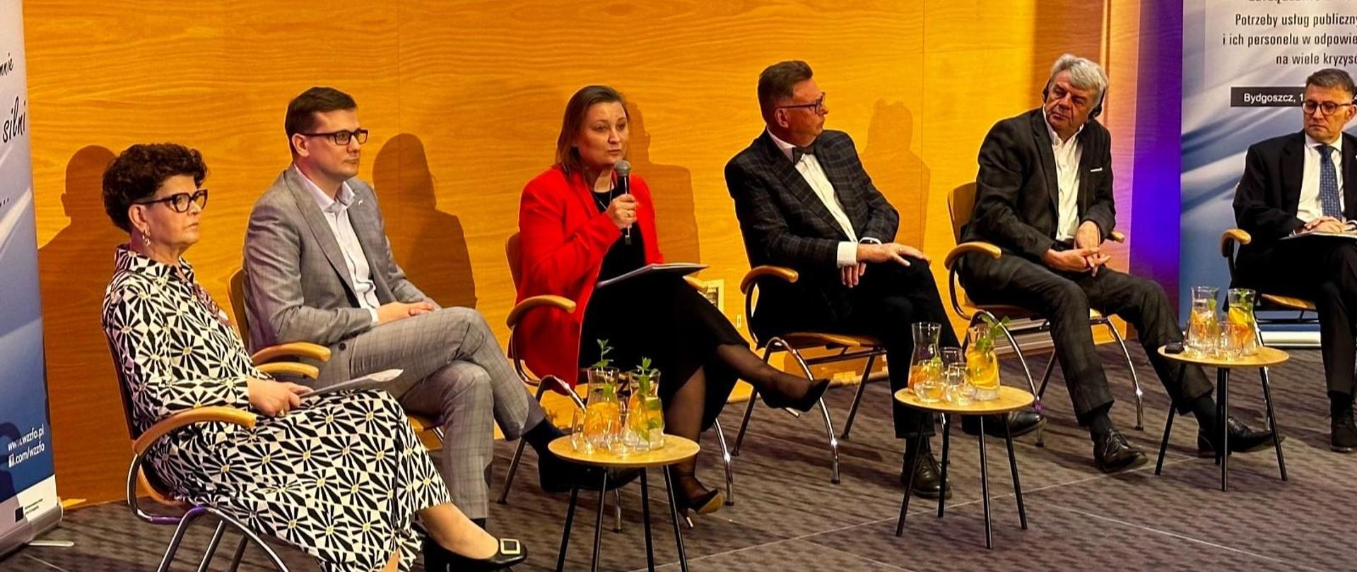 Na scenie na krzesłach siedzi sześć osób, dwie kobiety i czterech mężczyzn wśród nich wiceminister Piechna-Więckiewicz, która mówi do mikrofonu