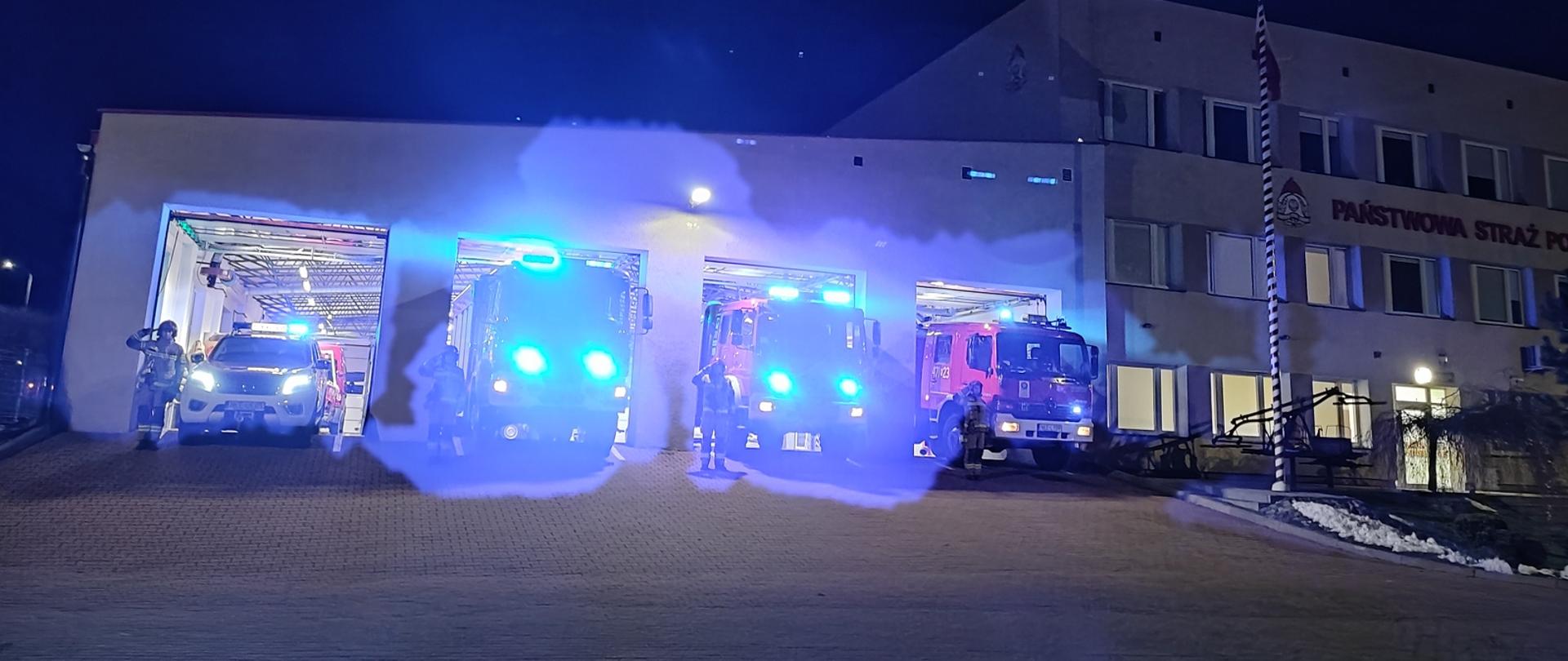 Zdjęcie wykonane porą wieczorową przed garażem JRG Kętrzyn. Czerwone samochody bojowe wystawione przed garaż z włączonymi niebieskimi światłami alarmowymi. Strażacy w piaskowych ubraniach bojowych oraz czerwonym hełmie stoją przy samochodzie oddają honor unosząc złączone dwa palce do hełmu. 