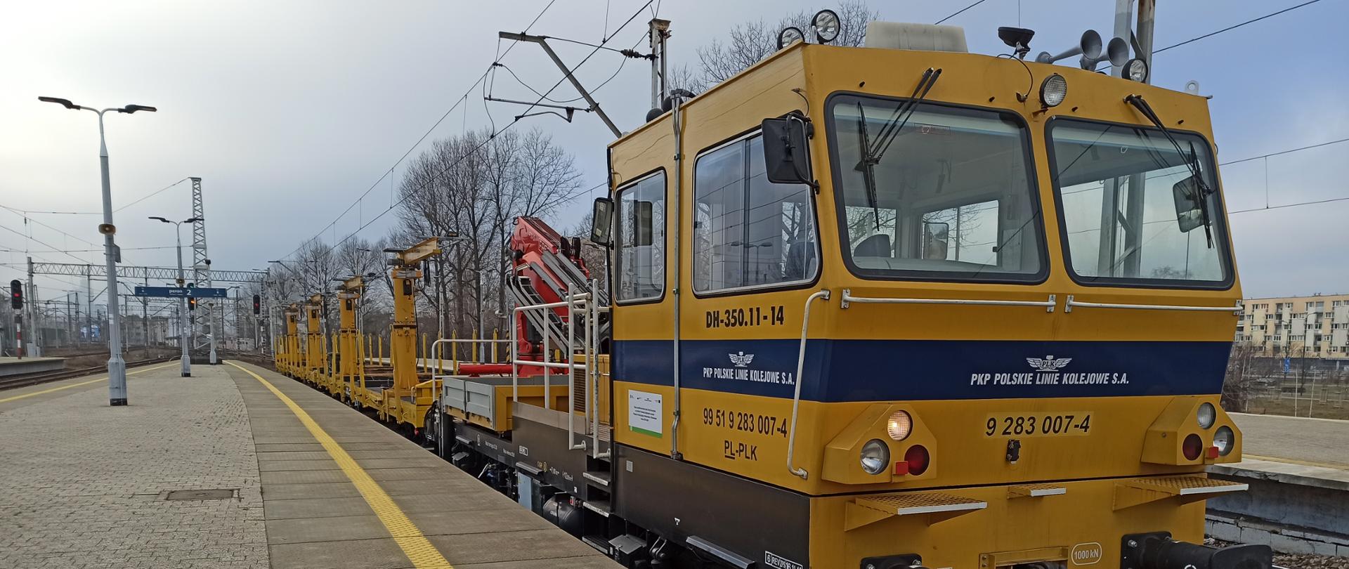 Zdjęcie z peronu kolejowego. Na torze stoi maszyna kolejowa, na której jest napis PKP Polskie Linie Kolejowe, oraz jest nakleja, mówiąca że sprzęt został zakupiony przy wsparciu funduszy unijnych. 