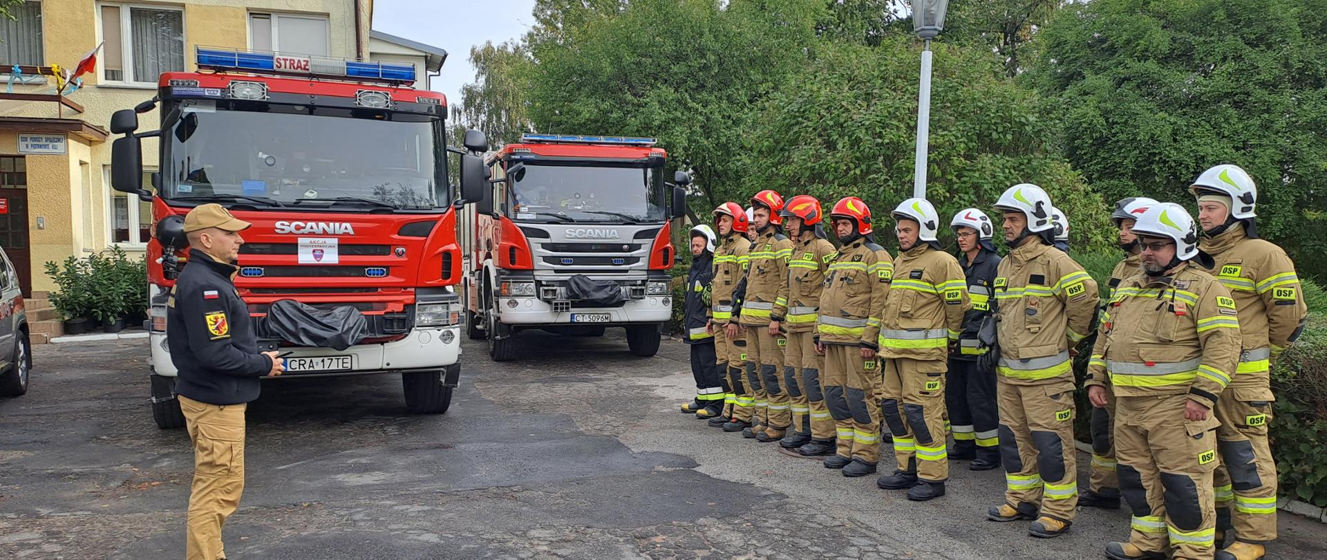 Zdjęcie przedstawia strażaków PSP oraz OSP podczas odprawy przed rozpoczęciem ćwiczeń obiektowych, na drugim planie wozy straży pożarnych.