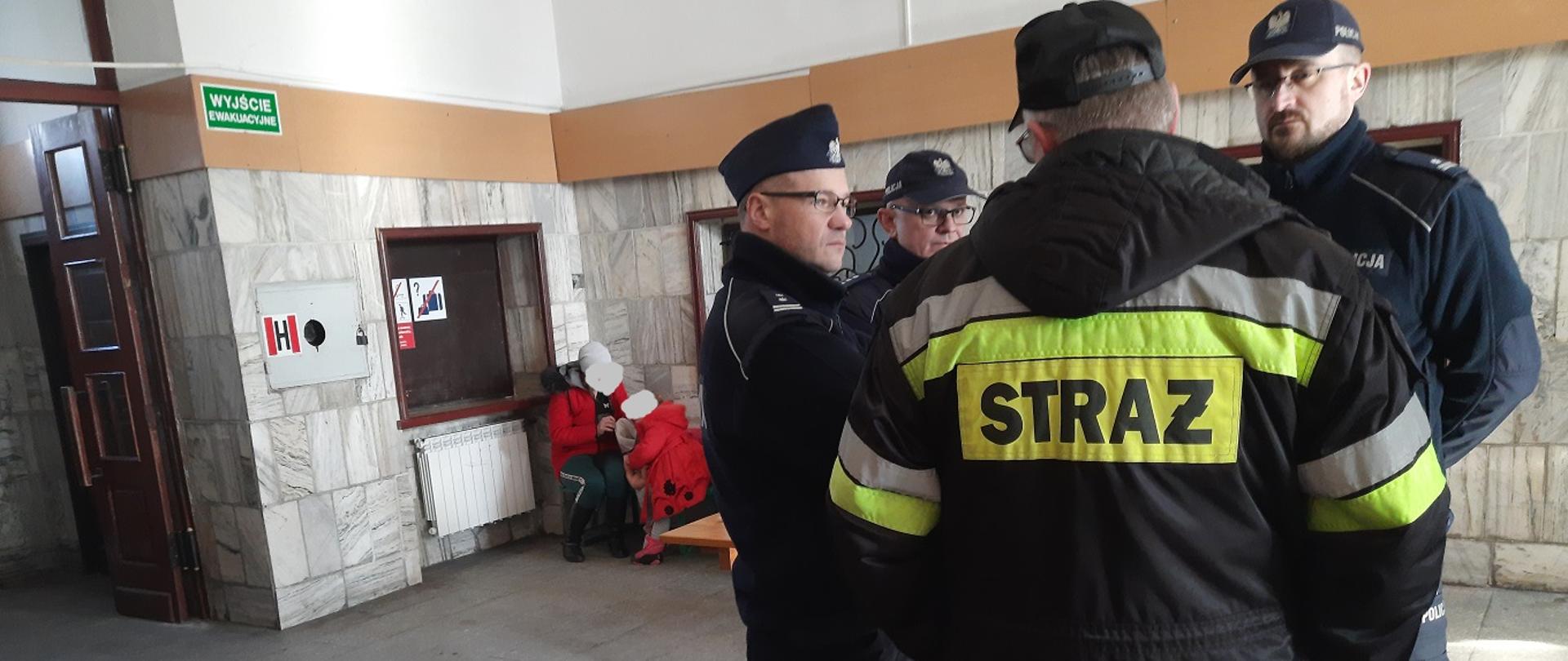 Zdjęcie obrazuje strażaka i 3 policjantów oraz dwie osoby cywilne w pomieszczeniu budynku
