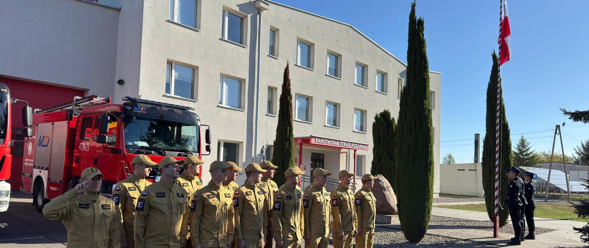 Moment podniesienia flagi państwowej na maszt. Strażacy stoją na baczność przed budynkiem komendy, przed samochodami bojowymi w piaskowych ubraniach koszarowych. Wszyscy spoglądają na wznoszącą się biało-czerwoną flagę. 