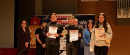 Pamiątkowe zdjęcie ze sceny po zakończeniu części konkursowej. Na pierwszym planie stoi troje młodych laureatów (dwóch chłopców i dziewczyna) trzymających dyplomy, a jeden z nich dodatkowo złotą statuetkę. W tle, za nimi, stoją dorośli – prawdopodobnie jurorzy lub organizatorzy. Sceneria to ta sama sala koncertowa z drewnianym wykończeniem i banerem „Jarosław 2026”. Atmosfera jest uroczysta i pełna dumy.
