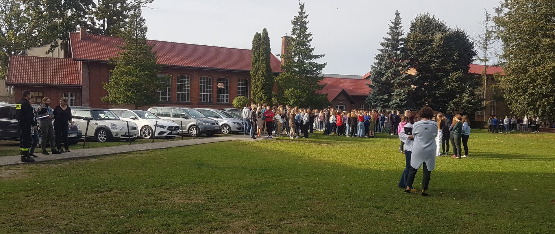 Próbne ewakuacje placówek oświatowych. Na zdjęciu uczniowie szkoły zebrani w miejscu zbiórki. Trwają ćwiczenia ewakuacyjne. Strażak wyznaczony do nadzorowania ćwiczeń ocenia przebieg wskazując uchybienia