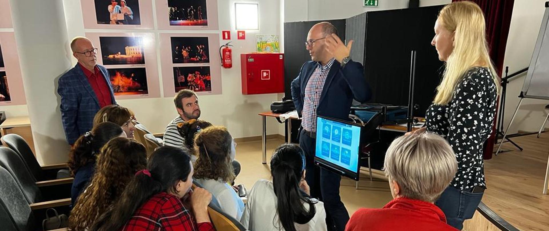 Kobieta i mężczyzna stoją w sali przy monitorze, mężczyzna coś wyjaśnia grupie osób zgromadzonych przed nimi.