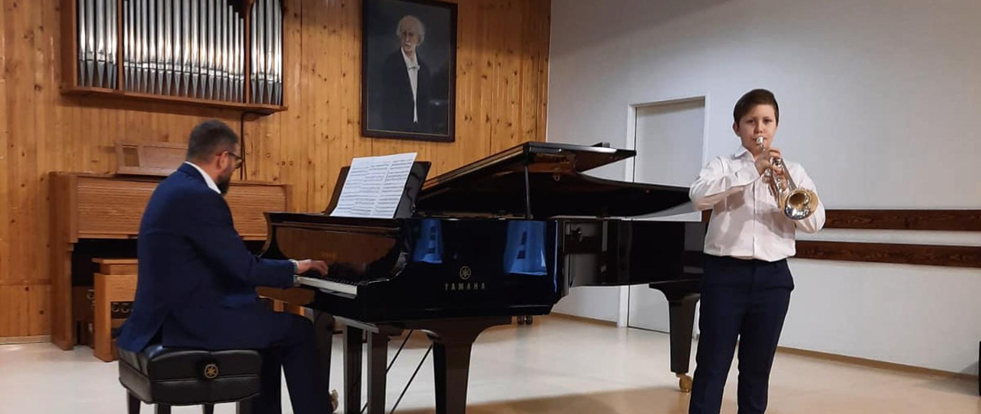 Chłopiec ubrany na galowo stoi i gra na trąbce, przy fortepianie siedzi pianista akompaniator, zdjęcie wykonane w sali koncertowej PSM w Koninie