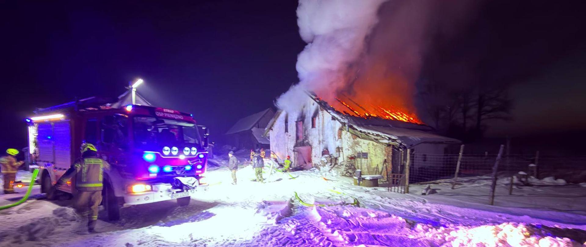 Zdjęcie zrobione nocą. Pali się budynek gospodarczy, widać płomienie na dachu i dużo białego dymu. Z lewej strony stoi samochód strażacki, przy nim strażacy.