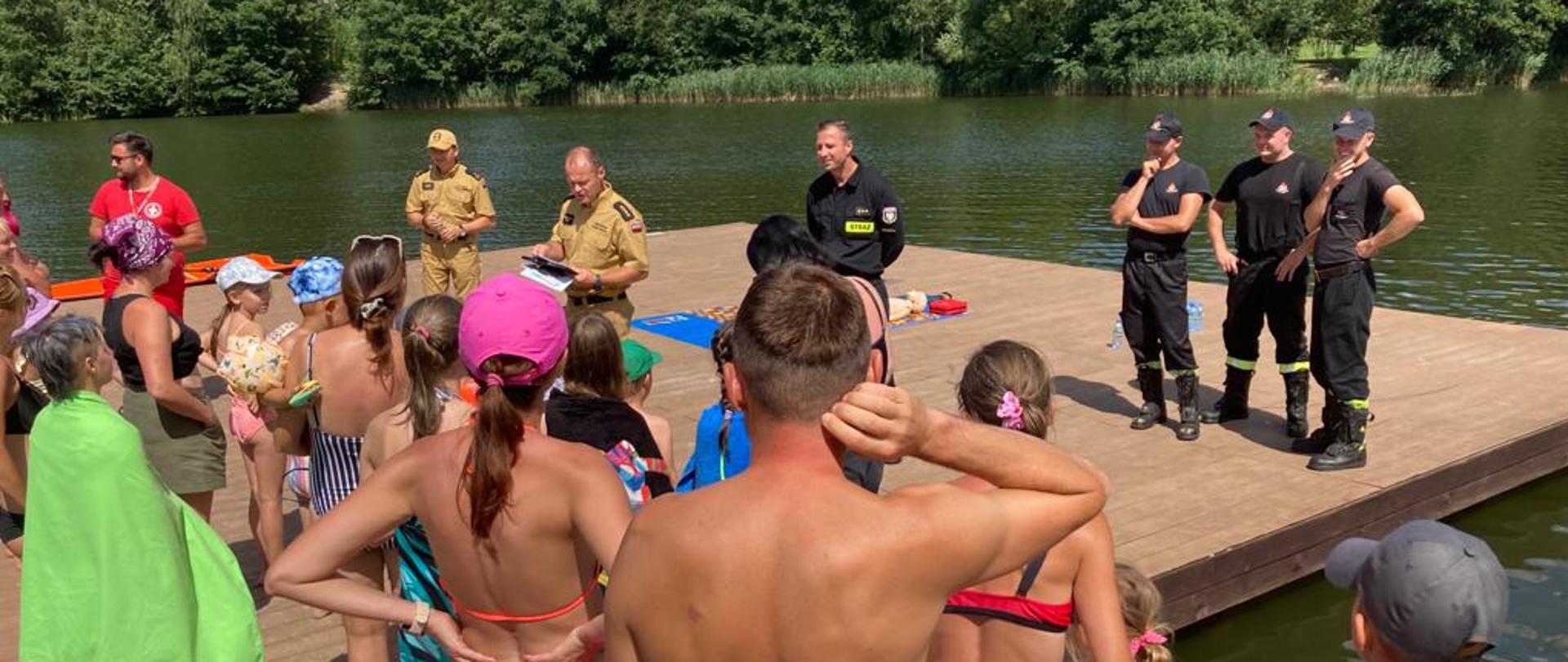 Na zdjęciu grupa ludzi stojących na pomoście słuchająca wykładu prowadzonego przez strażaków