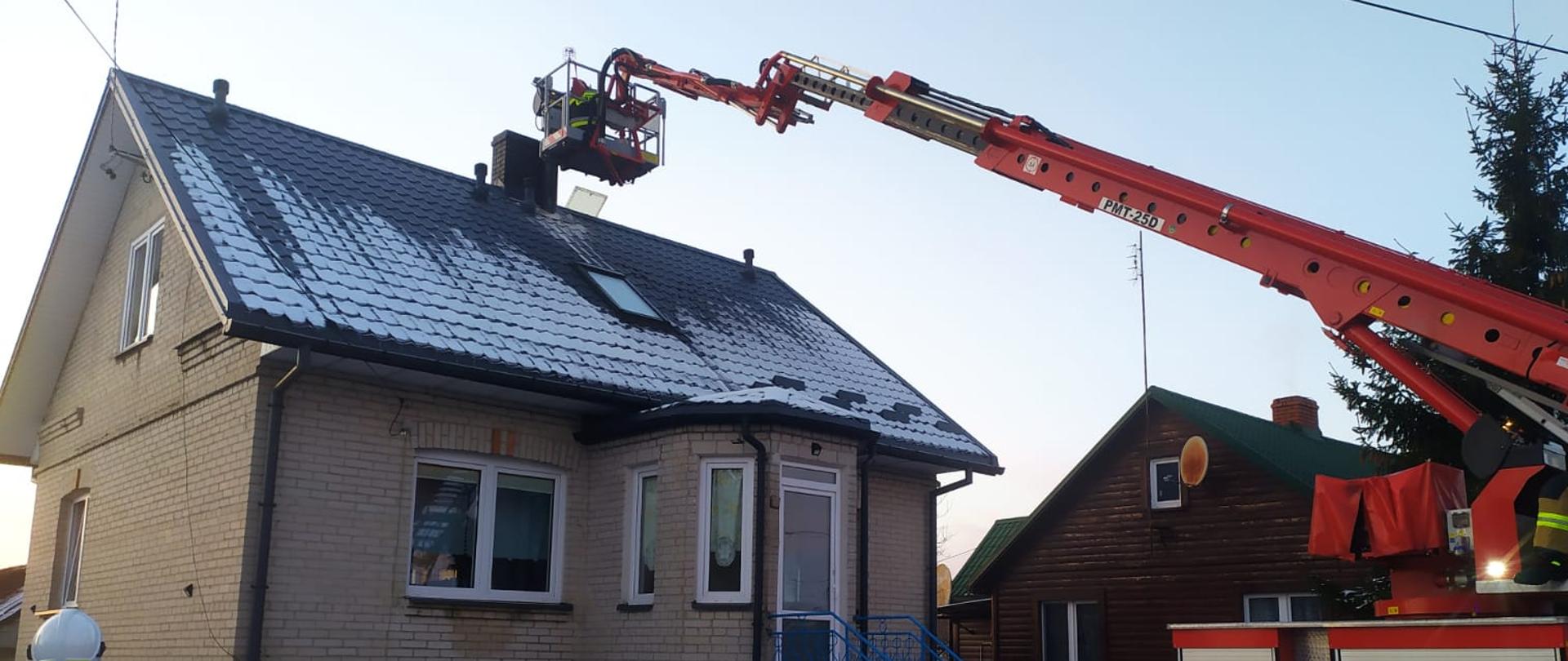 Ramię podnośnika hydraulicznego (pojazdu pożarniczego) rozstawione do komina budynku mieszkalnego. Budynek jednokondygnacyjny z poddaszem użytkowym z białej cegły, kryty grafitową blachodachówką. W koszu podnośnika pracuje strażak.