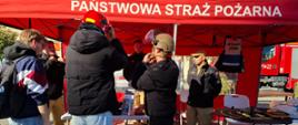 Zdjęcie przedstawia stoisko promocyjne Państwowej Straży Pożarnej pod czerwonym namiotem na miejskim placu. Dwóch młodych mężczyzn przymierza kaski ochronne – jeden czerwony strażacki, a drugi jasnobrązowy z podniesioną szybką. Przyglądają im się strażacy w mundurach oraz inny uczestnik wydarzenia. Na stoisku widoczne są materiały informacyjne, sprzęt strażacki oraz kamizelka z napisem „OPERATOR DRONA”.