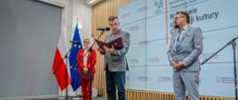 Zdjęcie z boku, na sali stoi minister Wieczorek, wiceminister Mrówczyńska w czerwonym garniturze i mężczyzna w jasnoszarym garniturze, minister trzyma w ręku czerwoną teczkę i czyta z niej do mikrofonu na stojaku.