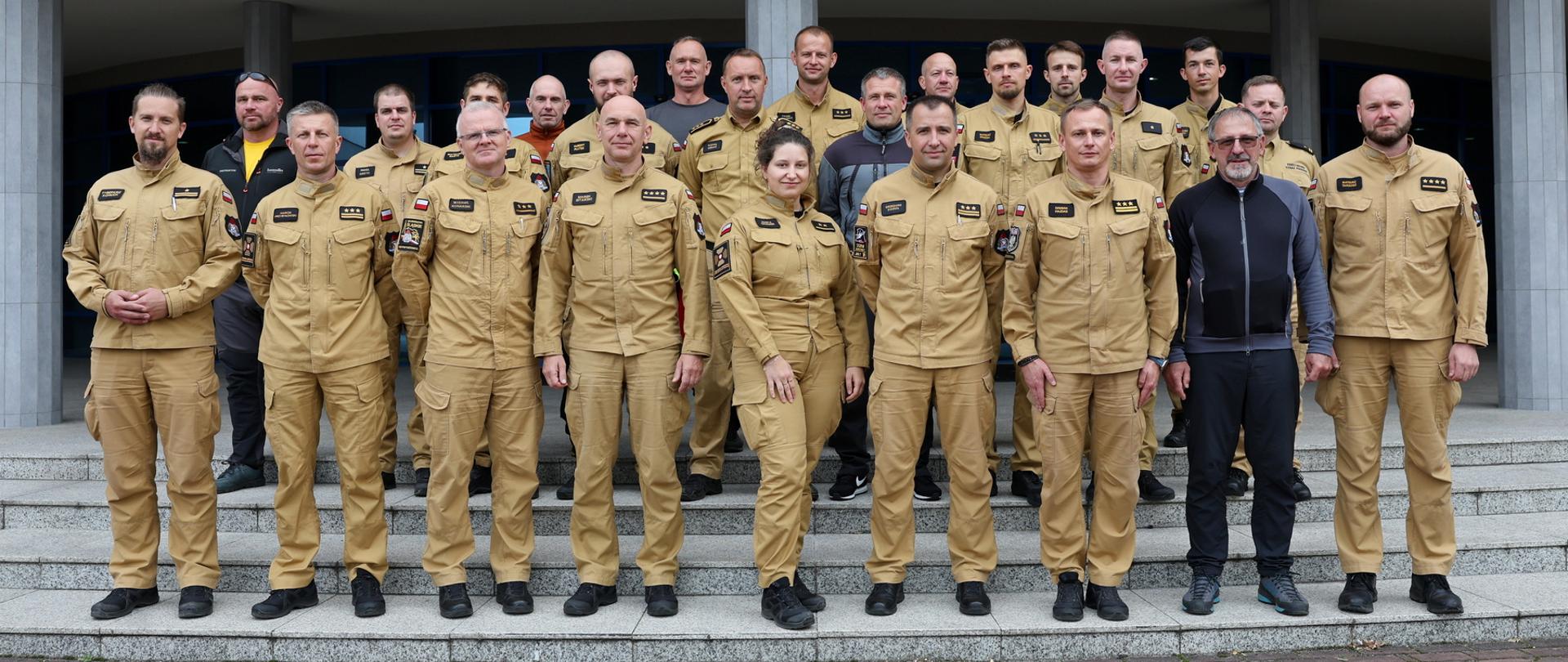 Pamiątkowe zdjęcie grupowe uczestników szkolenia kadry instruktorskiej ratownictwa wysokościowego KSRG stojących na schodach przed budynkiem. Większość osób ubrana jest w piaskowe mundury strażackie, pozostali w ciemną odzież