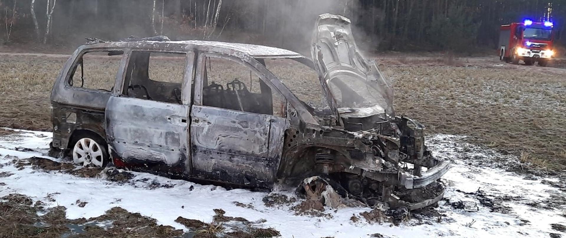 Na zdjęciu widoczny spalony wrak samochodu osobowego stojący w polu. Na drugim planie samochód gaśniczy straży pożarnej z włączonymi sygnałami błyskowymi.