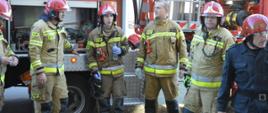 Pożar kamienicy na rawickim rynku. Na tle stojącego specjalnego samochodu pożarniczego z podnośnikiem hydraulicznym znajduje się sześciu strażaków uczestniczących w akcji. Pięciu z nich ma na sobie ubrania specjalne koloru piaskowego, szósty - czarny mundur dowódczo sztabowy. Strażak stojący w środku w lewej ręce trzyma butelkę z wodą.