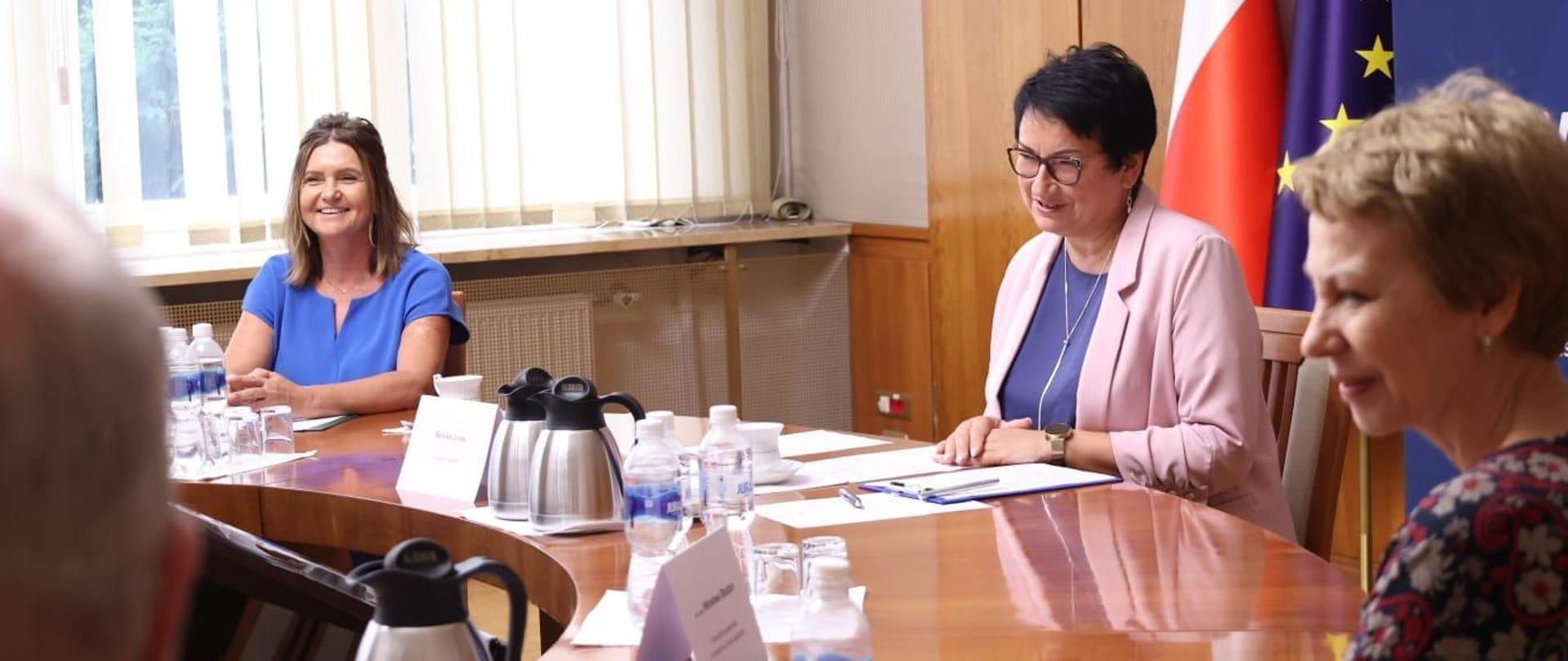 Wojewoda Opolski siedzi przy stole w Sali Złotej. Po lewej stronie zdjęcia siedzi Zastępca Dyrektora Wydziału Zdrowia. Na prawo siedzi jedna z powołanych konsultantek medycznych. Pani Wojewoda mówi coś, patrzy się na wprost. Wszyscy na zdjęciu się uśmiechają. 