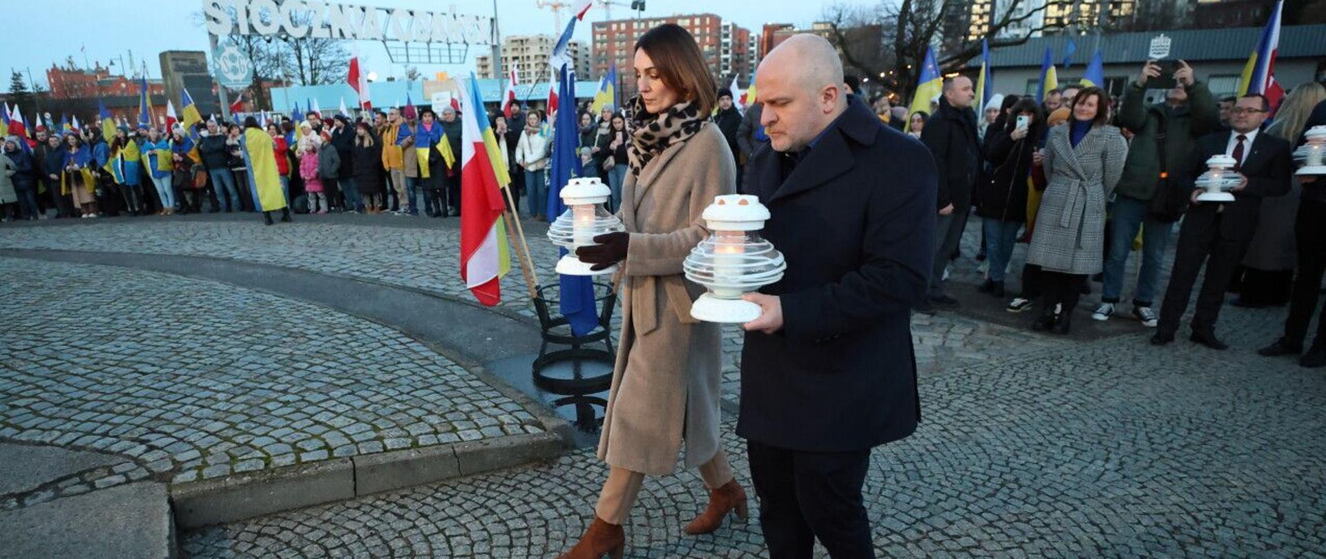 Na zdjęciu widać kobietę i mężczyznę, którzy trzymają znicze. W tle widać zgromadzenie ludzi