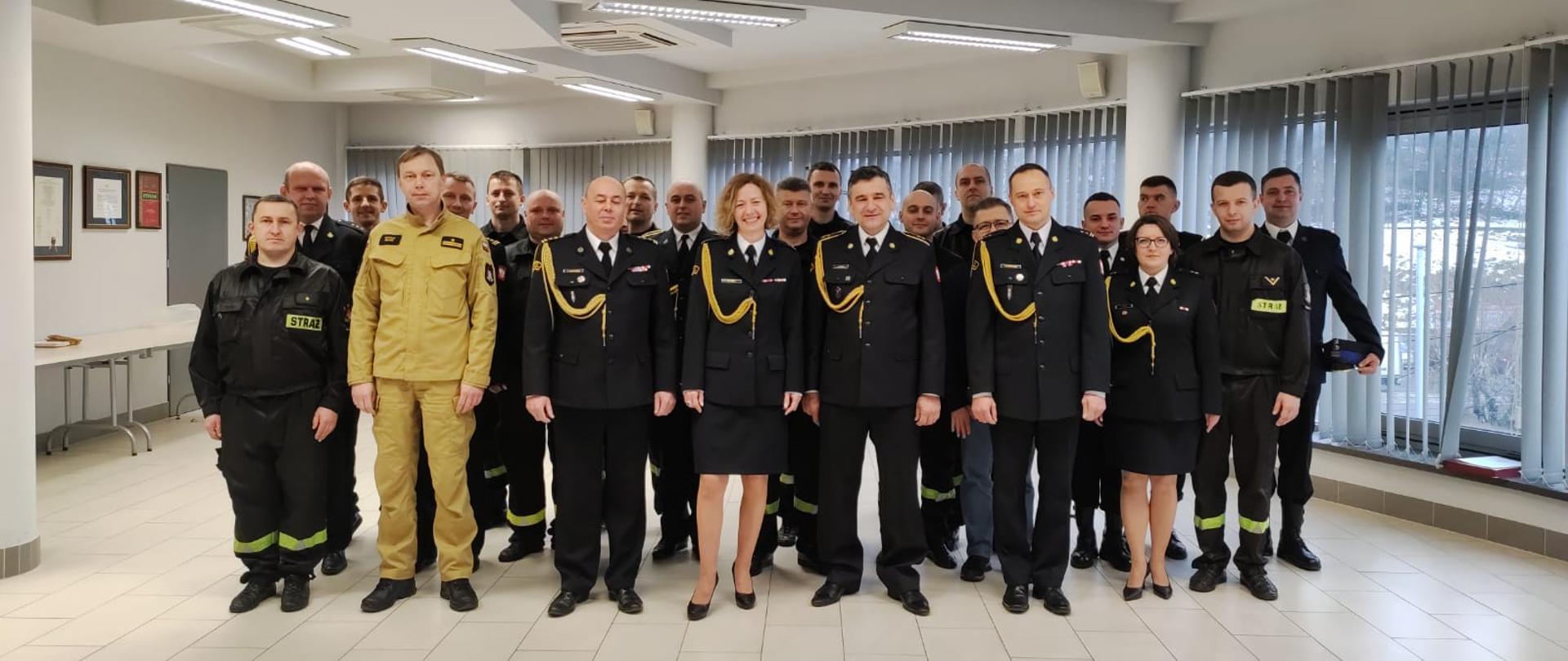 Kilkudziesięciu mężczyzn i kobiet ubranych w czarne ubrania wyjściowe, żółte sznury galowe, białe koszule, czarnym butami, stoją. W tle na ścianie sztandary w gablotach szklanych, żaluzje koloru szarego