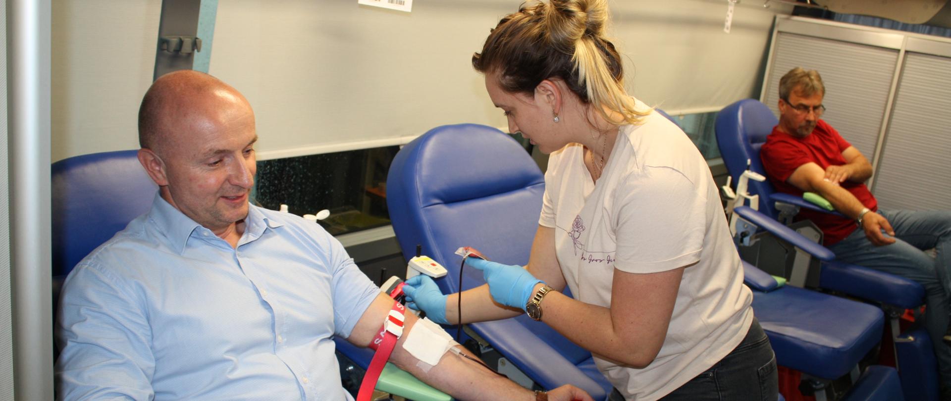 Na zdjęciu widać mężczyznę który przygotowuje się do oddania krwi podczas akcji „Ratujemy – dajemy”. Jest to komendant Miejski PSP w Krośnie. Mężczyzna oraz stojąca obok niego pielęgniarka znajdują się w autobusie, w którym osoby chętne mogą oddać krew (tzw. „krwiobusie”). W tle na fotelu widoczny jest drugi mężczyzna, który już oddał krew.