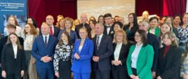 Zdjęcie zbiorowe, na sali pod ścianą stoi duża grupa ludzi i pozuje do zdjęcia wśród nich minister Nowacka.
