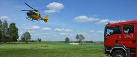 Start śmigłowca ratunkowego z pacjentem na pokładzie. Pacjent jest transportowany do szpitala. W tle drzewa, a z prawej strony wóz straży pożarnej, zabezpieczający operację. Na niebie chmury.