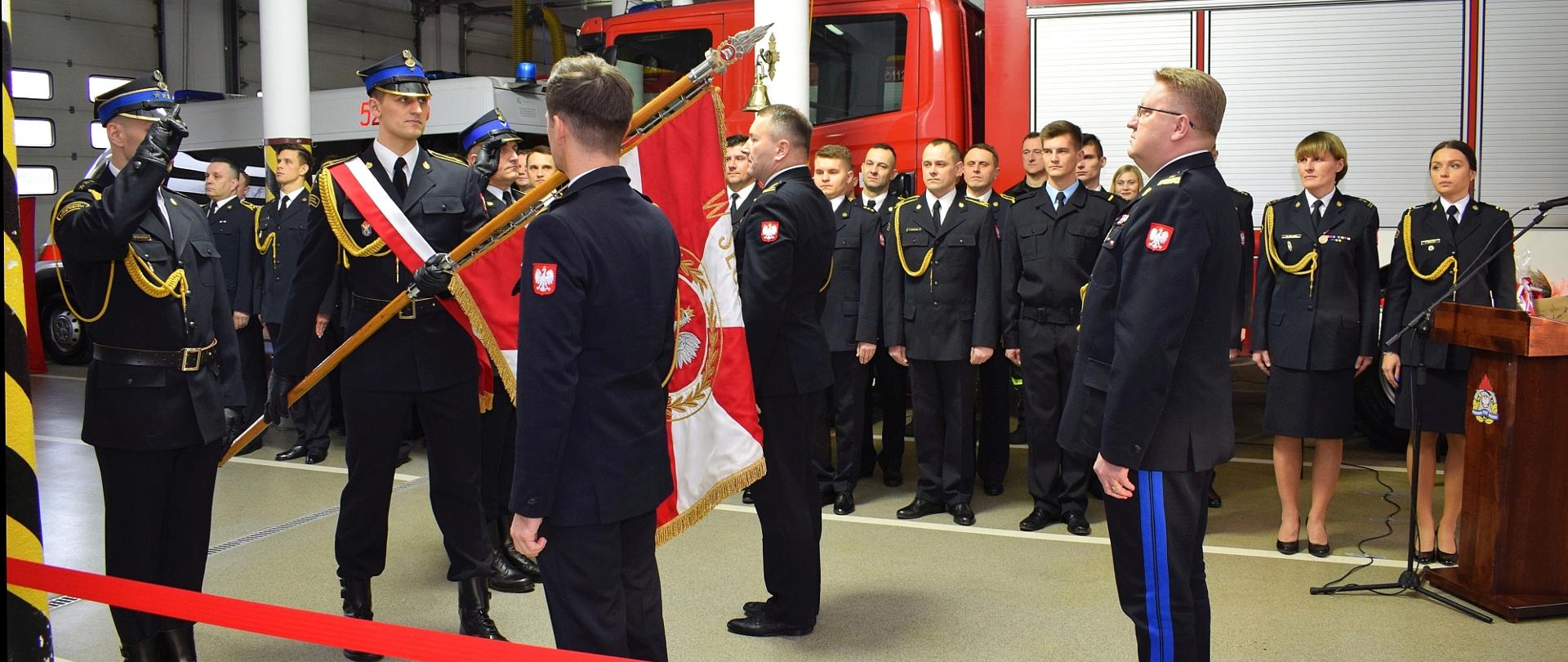 salutujący poczet sztandarowy komendy powiatowej PSP w Rawie Mazowieckiej, obok sztandaru stoją ustępujący i nowo powołany komendant powiatowy przed sztandarem stoi komendant wojewódzki PSP w Łodzi. w tle straży uczestniczący w zbiórce za nimi samochód pożarniczy