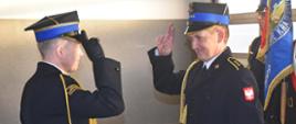 Zdjęcie wykonano w budynku Komendy Powiatowej Państwowej Straży Pożarnej w Sępólnie Krajeńskim w pomieszczeniu garaży. Komendant Powiatowy Państwowej Straży Pożarnej starszy brygadier Maciej Suchomski salutuje naprzeciw stojącemu aspirantowi sztabowemu Zenonowi Bingerowi. Oboje ubrani w mundury wyjściowe, na dalszym planie poczet sztandarowy. 