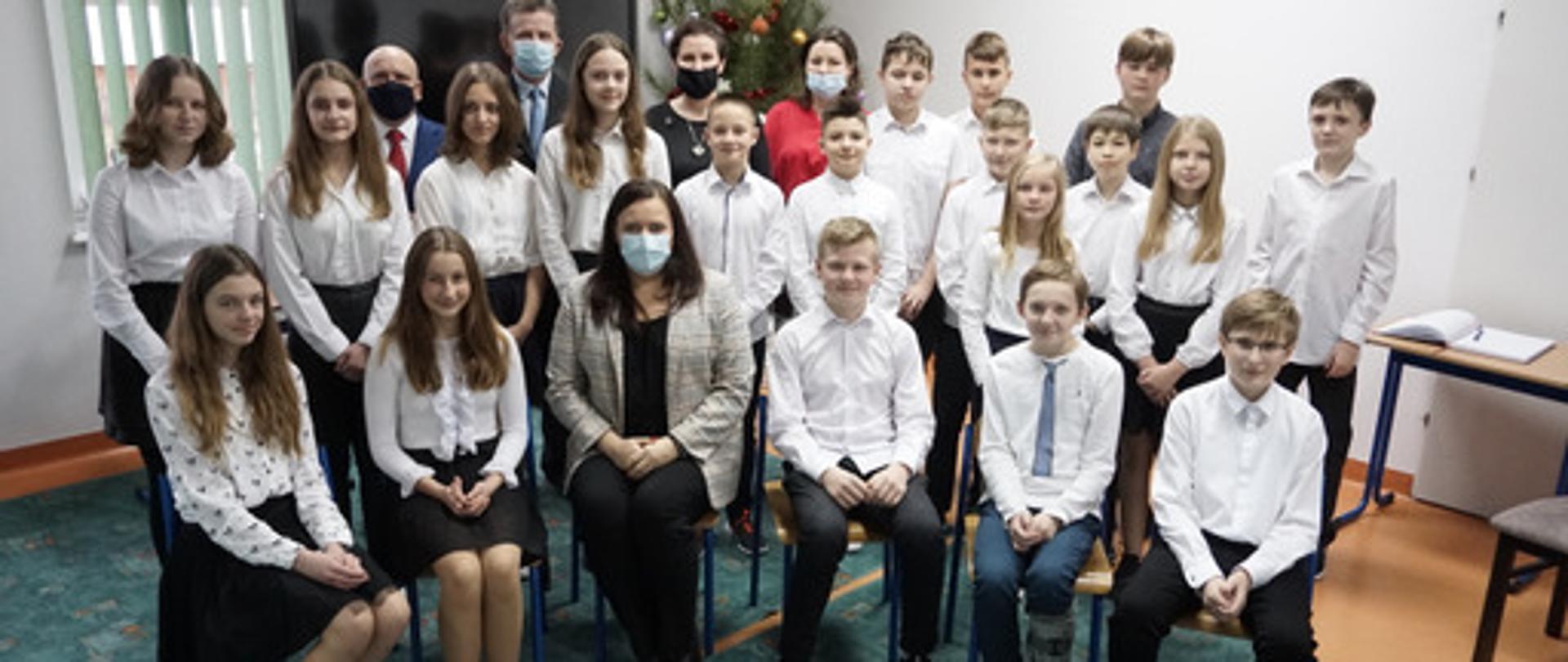 W grupie siedzą i stoją dzieci. Za nimi czwórka dorosłych. Pośrodku na krześle wiceminister Małgorzata Jarosińska-Jedynak.