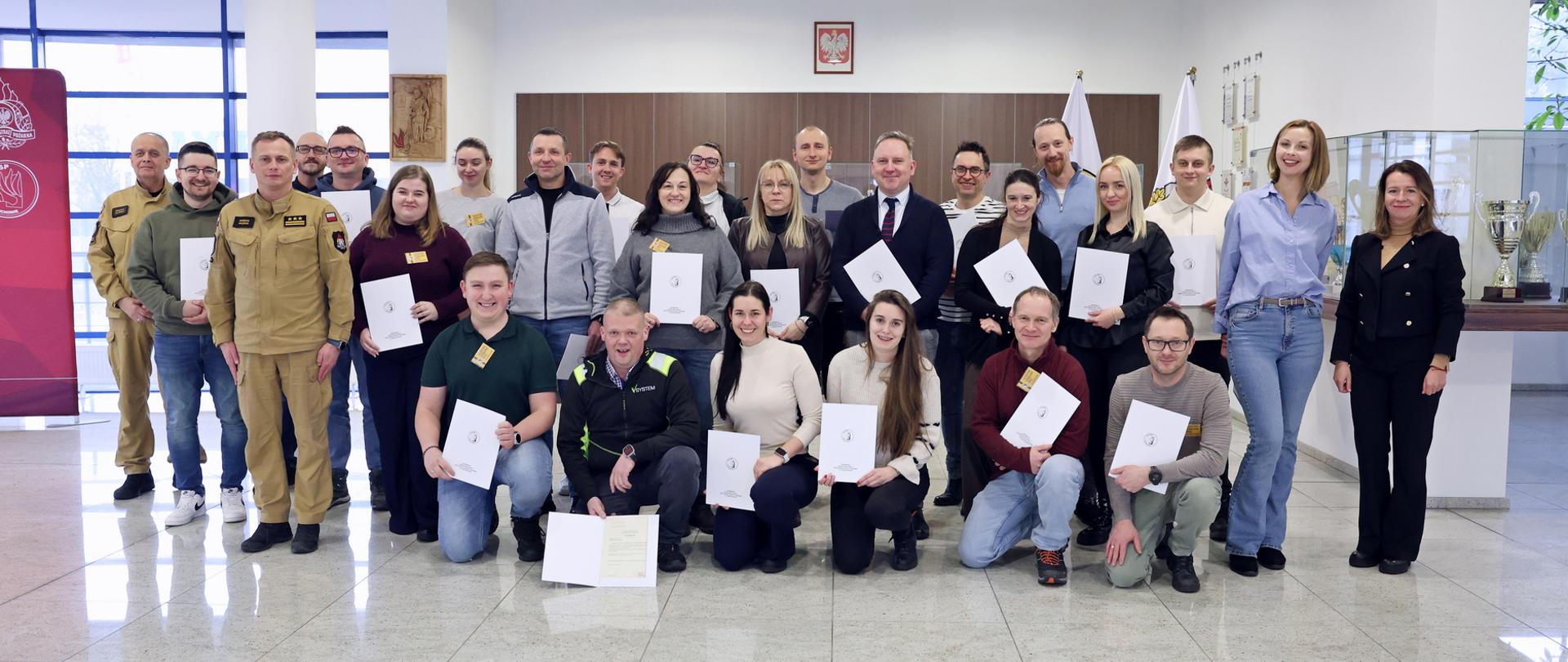 Pamiątkowe zdjęcie grupowe uczestników szkolenia inspektorów ochrony przeciwpożarowej w holu budynku. 23 osoby stoją w kilku rzędach, część trzyma dokumenty potwierdzające ukończenie szkolenia. Między uczestnikami szkolenia stoi dwóch funkcjonariuszy Państwowej Straży Pożarnej w piaskowych mundurach służbowych, w tym Zastępca Komendanta Centralnej Szkoły Państwowej Straży Pożarnej oraz kobieta. W tle widoczne są gabloty z pucharami oraz symbole państwowe