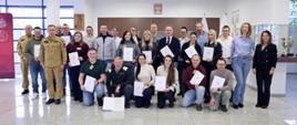 Pamiątkowe zdjęcie grupowe uczestników szkolenia inspektorów ochrony przeciwpożarowej w holu budynku. 23 osoby stoją w kilku rzędach, część trzyma dokumenty potwierdzające ukończenie szkolenia. Między uczestnikami szkolenia stoi dwóch funkcjonariuszy Państwowej Straży Pożarnej w piaskowych mundurach służbowych, w tym Zastępca Komendanta Centralnej Szkoły Państwowej Straży Pożarnej oraz kobieta. W tle widoczne są gabloty z pucharami oraz symbole państwowe