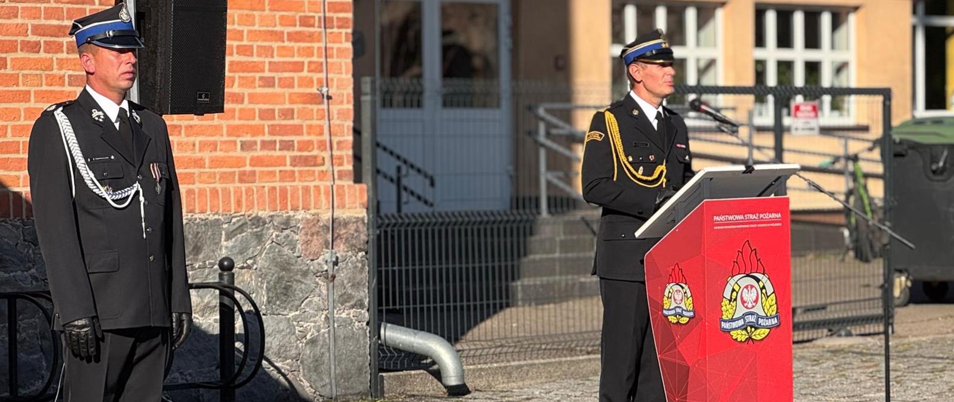 Na zdjęciu widzimy dwóch mężczyzn w mundurach strażackich stojących na placu przed budynkiem z czerwonej cegły. Po lewej stronie stoi mężczyzna w czarnym galowym mundurze z czapką z niebieskim otokiem. Ma białą ozdobną linkę na ramieniu oraz ciemne rękawiczki. Stoi na baczność, obok niego widać głośnik ustawiony na statywie. Po prawej stronie, za mównicą, stoi drugi mężczyzna również w czarnym mundurze, ale z ozdobnym złotym sznurem na ramieniu. Ma czapkę z niebieskim otokiem i przemawia do mikrofonu. Mównica jest czerwona, ozdobiona dużym herbem Państwowej Straży Pożarnej oraz napisem „Państwowa Straż Pożarna”. Za nimi widoczny jest budynek z oknami i ogrodzeniem, a na pierwszym planie widać brukowany plac, na którym odbywa się uroczystość.