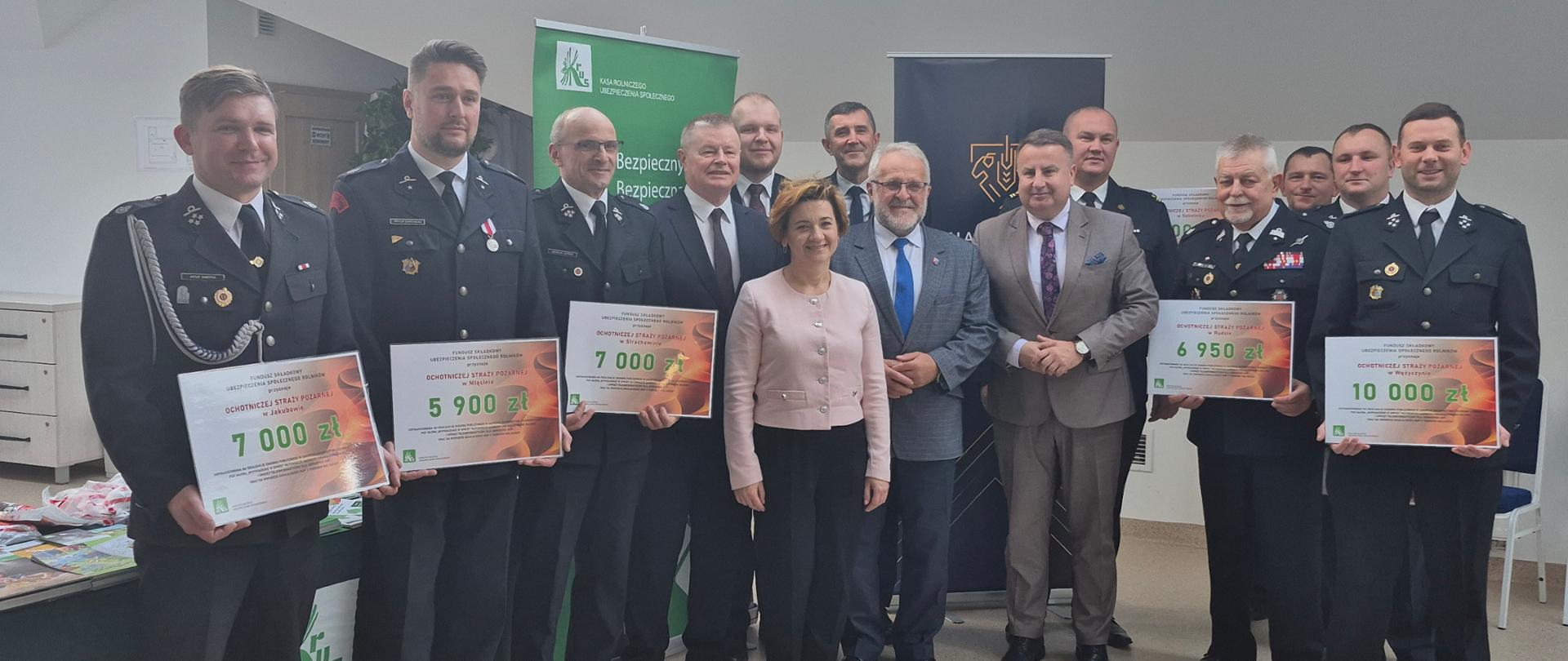 Jedna kobieta i trzynastu mężczyzn stoi tle banerów KRUS i Gminy Latowicz, część z nich w galowych mundurach strażaka, trzymają plansze z kwotą otrzymanego wsparcia.