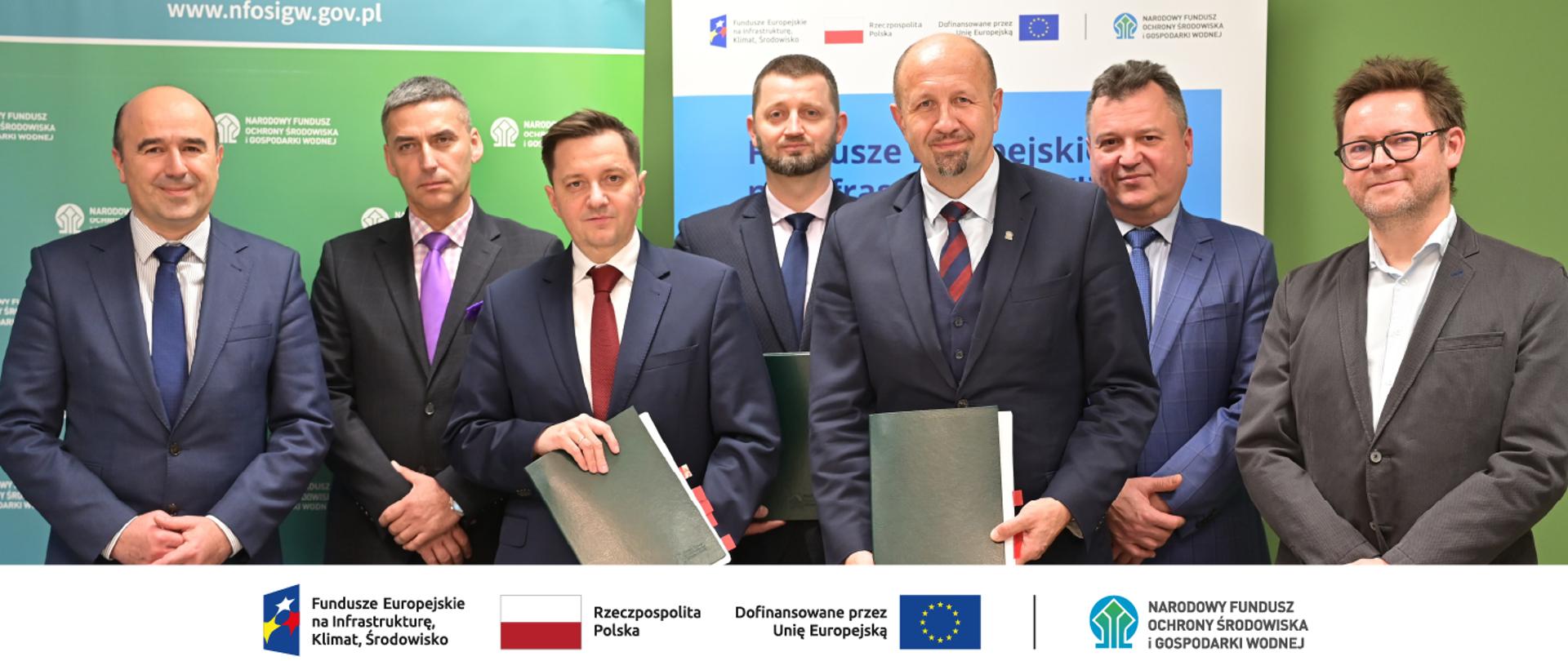 Zdjęcie grupowe. Stojący mężczyźni. Od lewej Stanisław Longawa, dyrektor Regionalnego Zarządu Gospodarki Wodnej we Wrocławiu, Tomasz Wujek NFOŚiGW, Mateusz Balcerowicz, prezes Państwowego Gospodarstwa Wodnego Wody Polskie, Jan Grygier, dyrektor Regionalnego Zarządu Gospodarki Wodnej w Gliwicach, Robert Gajda zastępca prezesa zarządu NFOŚiGW, Robert Markiewicz, dyrektor Departamentu Adaptacji do Zmian Klimatu NFOŚiGW, Krzysztof Witkowski NFOŚiGW. W stopce zdjęcia ciąg logotypów: Fundusze Europejskie na Infrastrukturę, Klimat, Środowisko, flaga Rzeczypospolita Polska, flaga UE i Dofinansowane przez UE, NFOŚiGW.