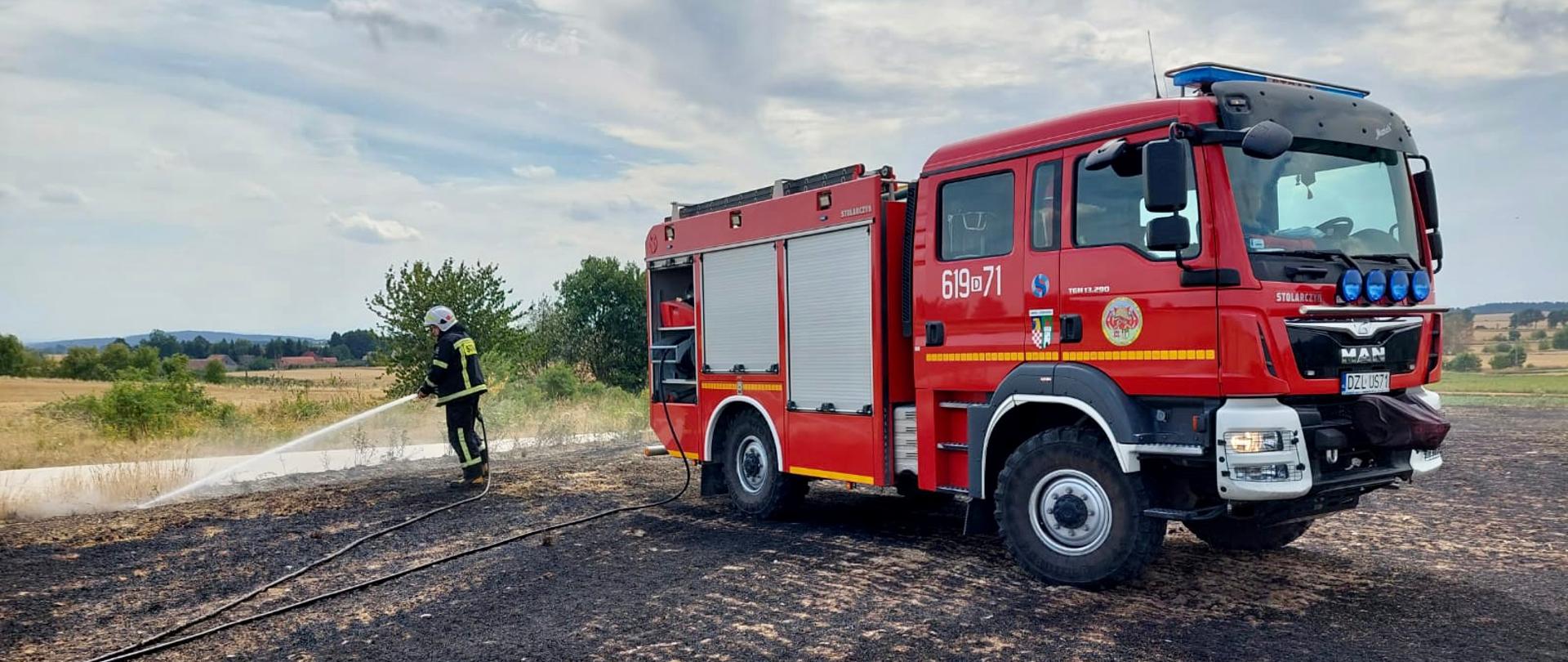 czerwony pojazd strażacki na wypalonym ściernisku, po lewej stronie strażak dogaszający linią gaśniczą pożar