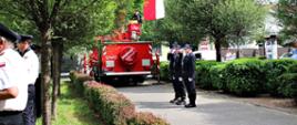 Plac Wolności w Rawiczu. Obok samochodu ratowniczego z podnośnikiem hydraulicznym stoi poczet flagowy wystawiony przez OSP w Izbicach. Druhowie oddają honor przez salutowanie. W tle budynki i drzewa.