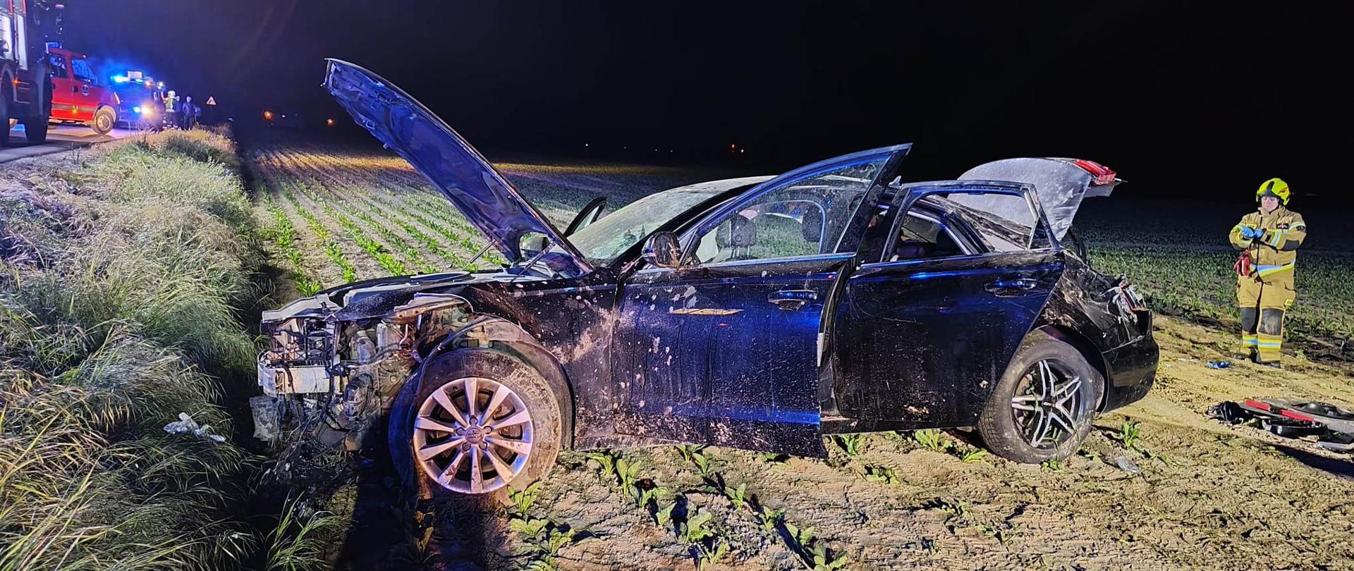 Uszkodzony pojazd na polu, jest ciemno, po prawej strażak
