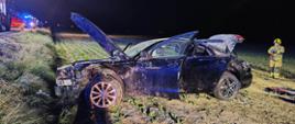 Uszkodzony pojazd na polu, jest ciemno, po prawej strażak