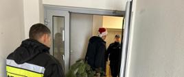 Policjant, mężczyzna w czapce Mikołaja oraz strażak w noszą choinkę do domu. Zdjęcie obrazuje moment przechodzenia przez drzwi wejściowe do budynku. 