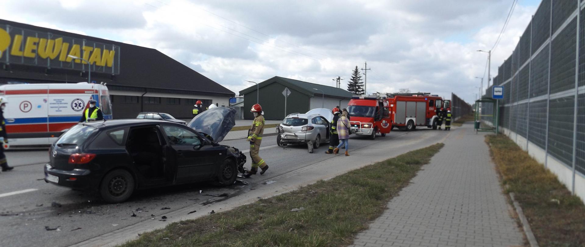 Zdjęcie przedstawia wypadek samochodowy – czarna mazda kombi z podniesioną maską, obok niej połamane plastiki i inne części. Przy samochodzie stoi strażak. Dalej widać uszkodzone srebrne Renault i samochody Straży Pożarnej. W tle stoi Pogotowie Ratunkowe.