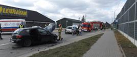 Zdjęcie przedstawia wypadek samochodowy – czarna mazda kombi z podniesioną maską, obok niej połamane plastiki i inne części. Przy samochodzie stoi strażak. Dalej widać uszkodzone srebrne Renault i samochody Straży Pożarnej. W tle stoi Pogotowie Ratunkowe.