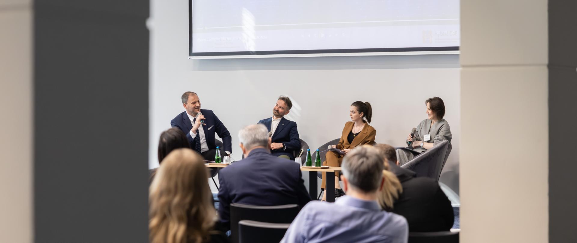 Panel dyskusyjny podczas konferencji – dwóch mężczyzn i dwie kobiety w formalnych strojach siedzą na scenie i rozmawiają, jeden z mężczyzn trzyma mikrofon; na widowni siedzą uczestnicy wydarzenia.