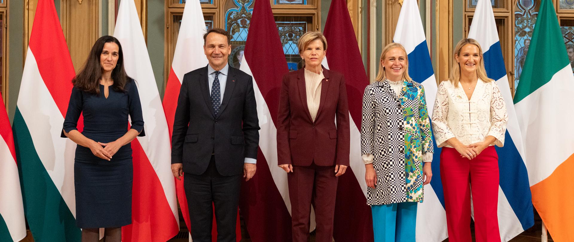 There are five people in the photo—four women and one man. They are looking straight ahead. Behind them are the flags of Hungary, Poland, Latvia, Finland, and Ireland.