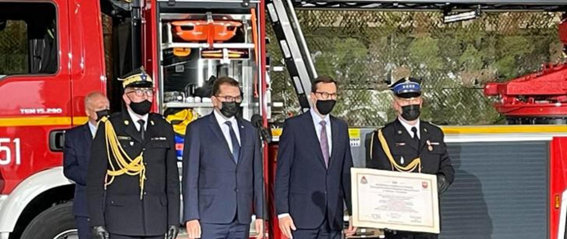  zastępca KG PSP, Wojewoda Małopolski, Premier RP oraz Komendant Powiatowy ustawieni w szeregu pozują do zdjęcia, komendant powiatowy trzyma w rękach akt przekazania, za nimi samochód ratowniczy PSP