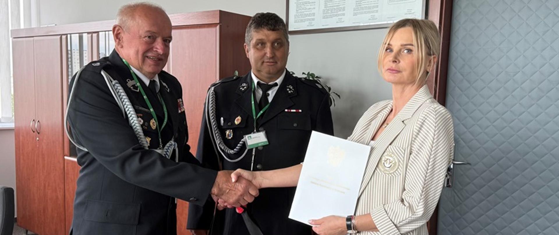 Na zdjęciu znajdują się kobieta w jasnym garniturze i dwóch strażaków w galowych mundurach. Strażak i kobieta podają sobie ręce, a między nimi widoczna jest czerwona torba ratownicza. Kobieta trzyma w ręku biały dokument z godłem Polski. W tle meble biurowe i tablica informacyjna na ścianie. 